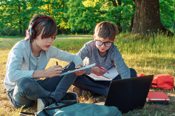 istock Boy in glasses carefully watching pupil in earphones who reading task from notebook, classmates study in park together. 1707451112