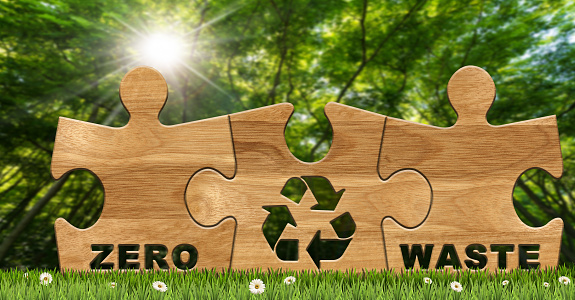 Close-up of three wooden puzzle pieces with text, Zero Waste, and a Recycling Symbol. On a green meadow with daisy flowers and a green forest on background. Sustainable Resources concept.