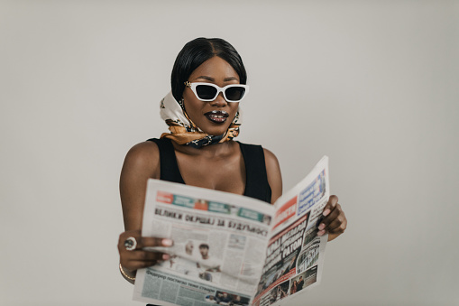 Portrait of a beautiful young black woman wearing an elegant black dress and headscarf reading newspapers, studio shot in front of a white background