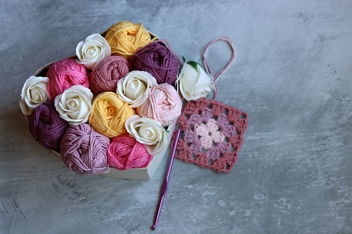 Multicolored balls of cotton yarn and white roses in a basket on a gray background with space for text. Handmade gift box with organic thread. Creative gift for crochet lover.