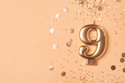 Gold candle in the form of number nine on peach background with confetti.