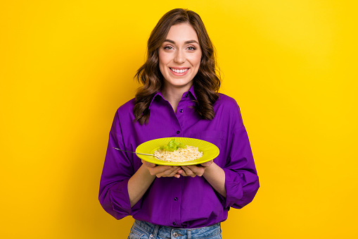 Portrait of cheerful lovely lady toothy smile arms hold showing noodles plate isolated on yellow color background.