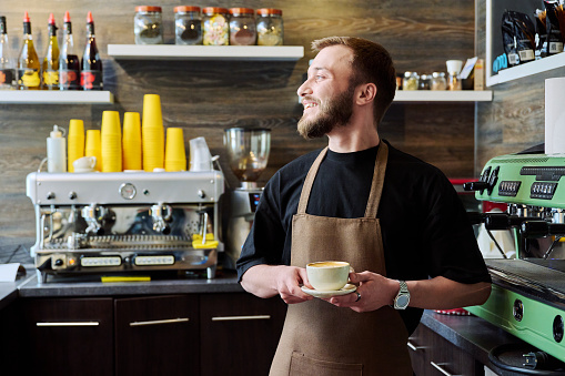 Portrait of young barista male in an apron with freshly prepared cappuccino coffee, standing behind counter in coffee shop, machine background. Small business service, coffee shop, staff work concept