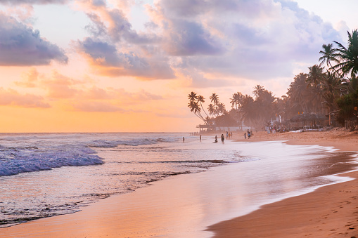 A beautiful sunset on a sandy beach with palm trees and people. The beach is located in Hikkaduwa. The water is with white foam, reflecting the orange hues of the sky. The sky is filled with scattered clouds that create a dramatic contrast with the sun. The beach is lined with palm trees that sway in the breeze. There are a few people on the shore, enjoying the view or walking along the sand. The mood of the photo is peaceful and serene, capturing the essence of a relaxing vacation.