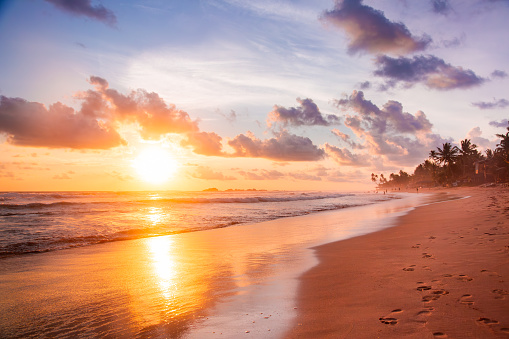 A stunning sunrise over a tropical beach with clouds and palm trees. The sun is rising over the horizon and is partially obscured by clouds, creating a dramatic effect. The sky is a gradient of orange and blue, with scattered clouds adding texture and depth. The beach is sandy and the water is a light blue, reflecting the colors of the sky. The waves are creating a peaceful atmosphere. There are palm trees on the right side of the image. The image has a warm and peaceful mood, conveying the beauty of nature and the start of a new day.