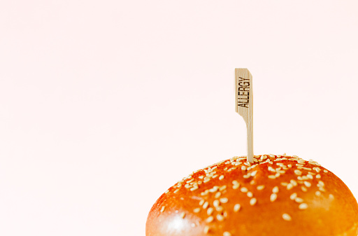 Close up of brioche bread buns topped with sesame seeds. Through the buns is a wooden stick marked with the word 'allergy'. Focus on the buns and stick with a white background.