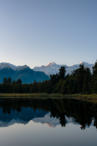 Dawn breaks over spectacular Lake Matheson in New Zealand, whilst beyond we see the snow capped peaks of the Southern Alps.