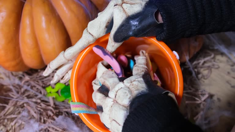 Skeleton bone hands showing colorful sweets in bucket and collects candy for trick or treat, orange pumpkin,