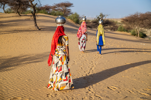 Indian young girls crossing sand dunes and carrying on their heads water from local well, Thar Desert, Rajasthan, India. Rajasthani women and children often walk long distances through the desert to bring back jugs of water that they carry on their heads.