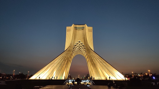 The mesmerizing photograph showcases the striking Azadi Tower, a majestic, towering structure adorned in pristine white. Its circular base and soaring, pointed pinnacle captivate the viewer's attention. A vibrant plaza surrounds the magnificent monument, which attracts a flurry of individuals