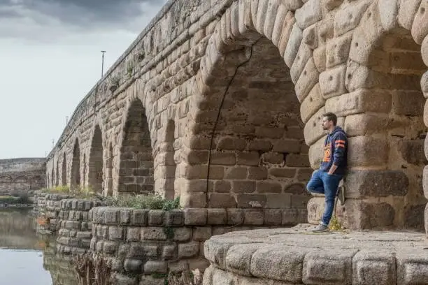 Photo of Young man in jeans and sweatshirt on the Roman bridge in Merida.