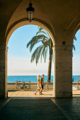 Wide shot of a couple on holiday walking under an archway on the Promenade des Anglais in Nice, France