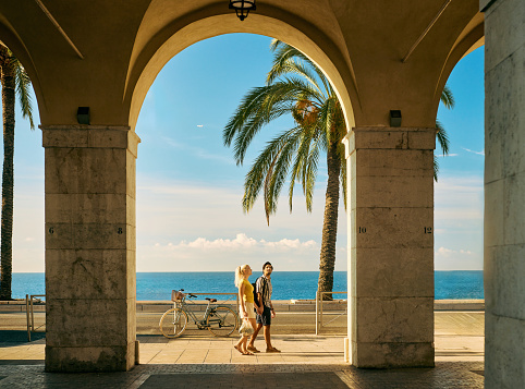 Wide shot of a couple on holiday walking under an archway on the Promenade des Anglais in Nice, France