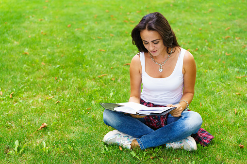 Smiling female student sitting on grass with notepad in hand, preparing for exams. Education and remote work concept. Soft selective focus. Caucasian girl with a pensive look