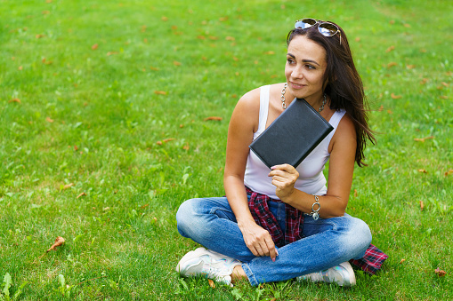 Smiling female student sitting on grass with notepad in hand, preparing for exams. Education and remote work concept. Soft selective focus. Caucasian girl with a pensive look