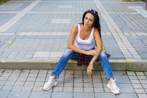 Happy young fashion woman sitting on the street sidewalk stylish female model in a white t-shirt and in dark blue jeans arms wide apart from joy