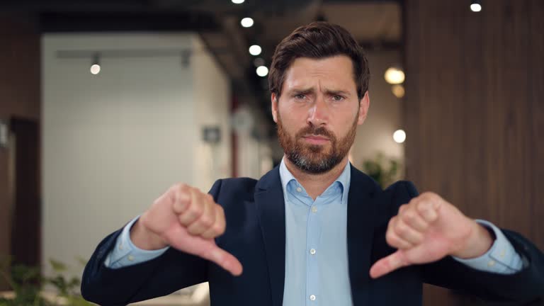 Portrait of nervous bearded caucasian man showing thumbs down and expressing negative emotions. Dissatisfied male feeling disappointment and injustice over background of personal designer cabinet.