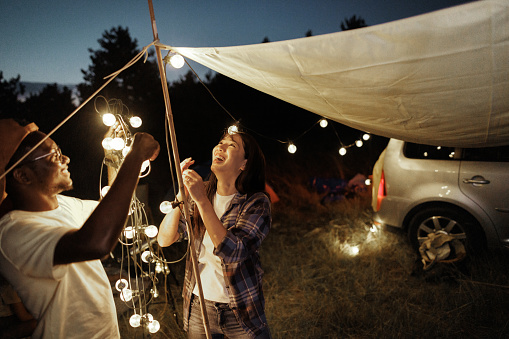 Young couple setting up string lights to decorate and lighten their camping spot at night