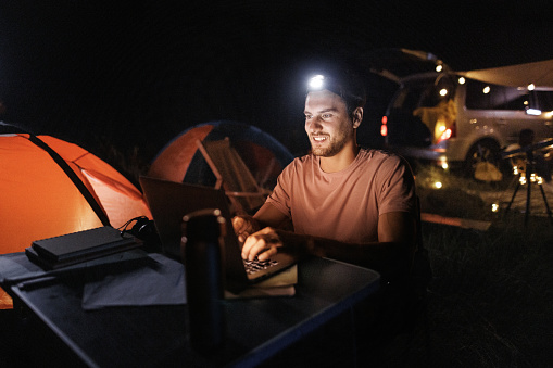 Young handsome casually clothed man working on laptop with headlamp on his head while camping in forest at night