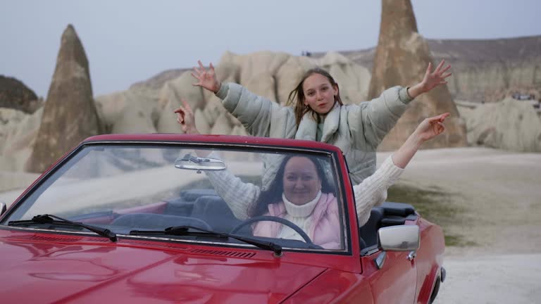 Happy mom daughter in cabriolet car wave hands look at camera in rocky desert.