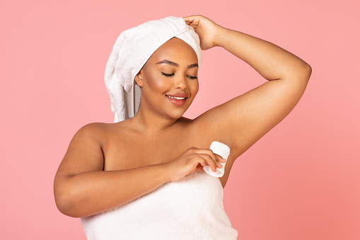 African American Plus Size Woman Applying Antiperspirant Stick On Armpit Caring For Herself Posing Wrapped In Towel Over Pink Studio Background. Sweat Protection And Freshness Concept