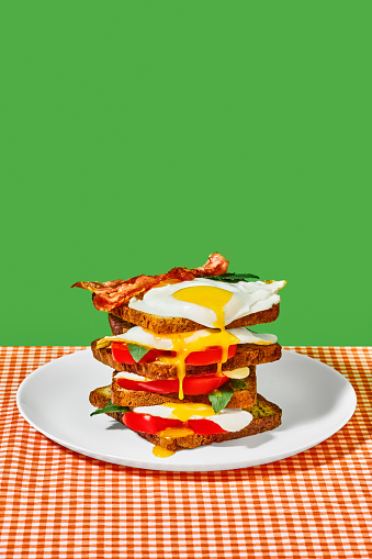 Toast bread, fiend eggs, bacon, tomatoes and basil on plate on checkered tablecloth over green background. Concept of breakfast, food, taste, creativity. Pop art photography. Poster. Copy space for ad