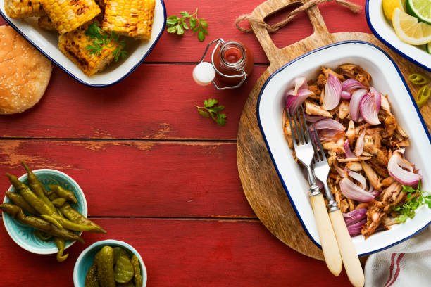 Pulled bbq chicken with baked onions on serving platter, bbq corn, pickles, chili peppers and buns for hot dogs and burgers, tomato sauce. Traditional American grilled summer bbq sandwiches dishes. pollo a la barbacoa desmenuzado con cebollas al horno en el plato para servir, maíz a la barbacoa, pepinillos, chiles y bollos para perros calientes y hamburguesas, salsa de tomate. platos tradicionales de sándwiches de barbacoa de verano a la parrilla a