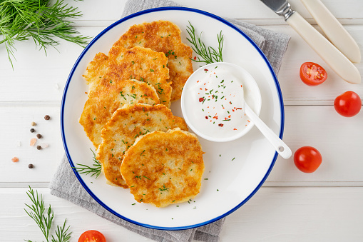 Potato pancakes, latkes or draniki with fresh herbs and sour cream on a plate on a white wooden background. Top view, copy space