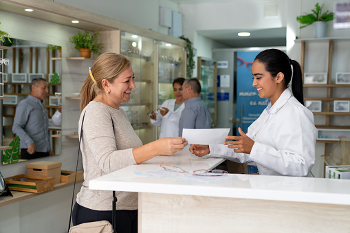 Happy Latin American woman showing prescription to the eye doctor while buying glasses at the optician's shop