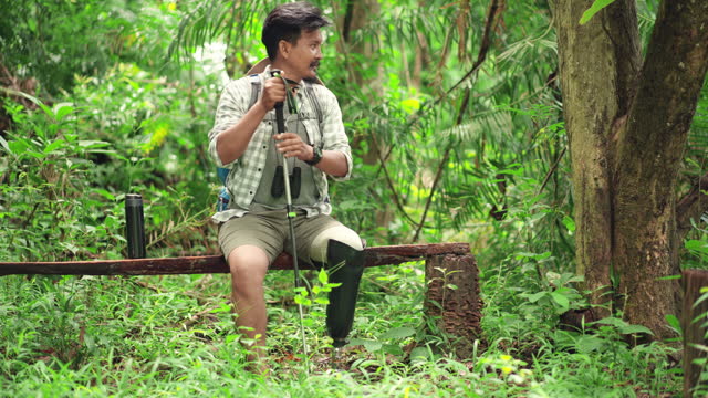 Mid adult disabled Asian man sitting on a wood bench in the forest for relaxation after hiking for a long time, male is wearing prosthetic leg in order to go forest hiking to his next destination and using hiking pole for helping him during walking