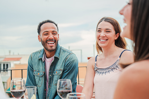 Smiling man laughing with two women friends celebrating a lunch party with wine glasses, sitting at table in the restaurant terrace. Group of three people relaxing and having a great time together. High quality photo