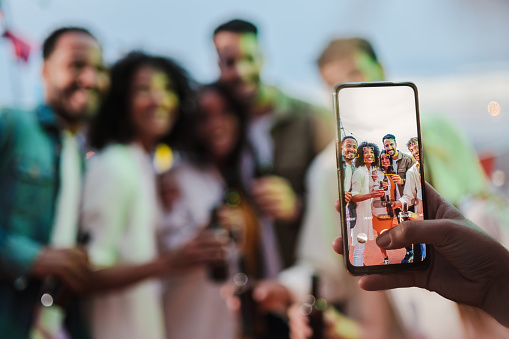 Focused on the cellphone screen. Blurred group of a young adult friends shooting a picture with a smart phone on a social gathering party celebration, having fun together. blur technique. High quality photo