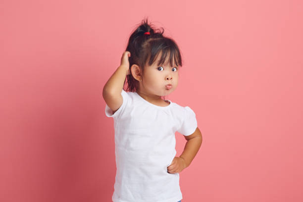 Little funny girl pouts lips and scratching her head with a thoughtful expression, has a puzzled look, wearing in white neutral t shirt standing over pink isolated baclground.