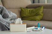 Cold medicine on the background of a sofa with pillows and a blanket. Tablets, spray and thermometer are on the table