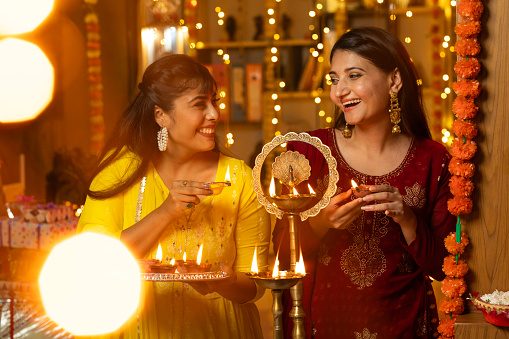 Beautiful young sisters looking at each other and smiling while lighting diyas at home during Diwali celebration