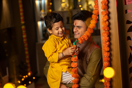 Smiling father carrying cute son playing with floral garden hanging on wooden wall at home during Diwali festival