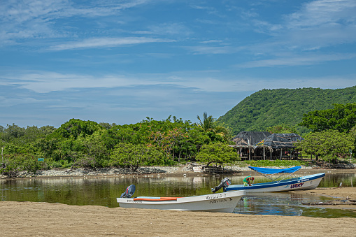 Zihuatanejo, Mexico - July 18, 2023: Playa Larga. at the channel to Laguna de Potosi, 2 small fishing boats with green mountain and jungle in back under blue cloudscape