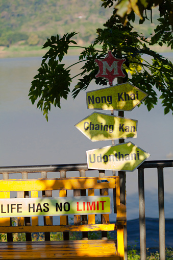 Yellow wooden directional signs to cities at Mekong river in north Thailand. Sign is placed near a tree on promenade with small market and retail area and boat piers. On signs are names of cities Chiang Khan, Udon Thani. Ning Kai. Below is text LIFE HAS NO LIMITS. Place is east of Chiang Khan