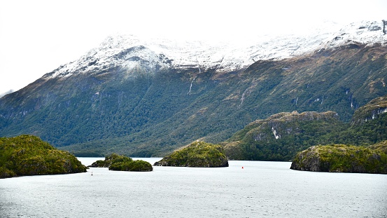 Snow topped mountains rise out of the waters of the fjords of Chile beyond a narrow channel.