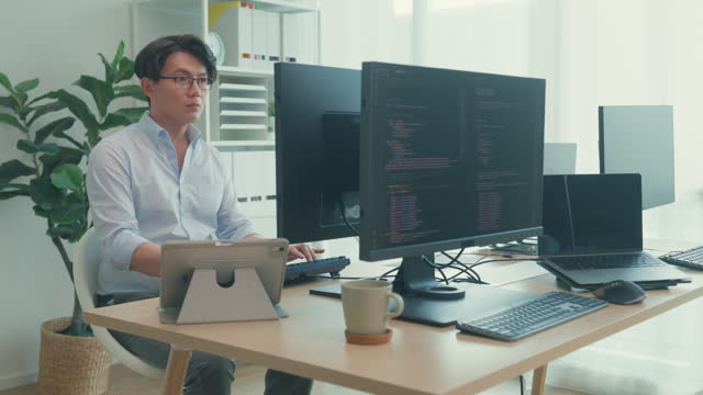 Young Asian man software developers using computer to write code sitting at desk with multiple screens work at office. Programmer development.