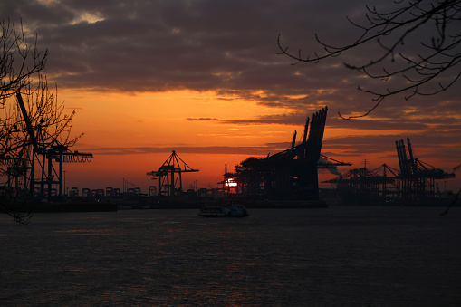 Cranes at the port of Hamburg at sunset, Germany.  Port of Hamburg is a sea port on the river Elbe in Hamburg, Germany, 110 kilometres from its mouth on the North Sea. It is Germany's largest port. Hamburg is the second-busiest port in Europe (after Rotterdam) and 15th-largest worldwide.
