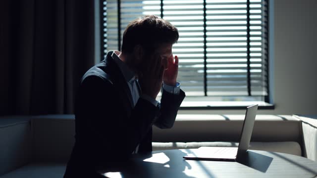 Side view of entrepreneur conducting business correspondence with partners online via wireless computer in stylish office. Focused man feeling sudden sharp headache, closing eyes and touching head.