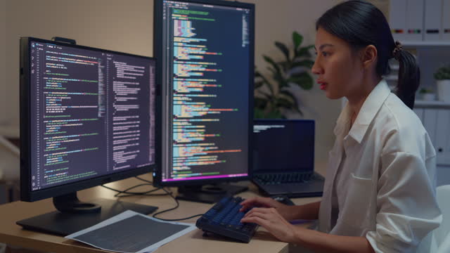 Young Asian woman software developers using computer to write code sitting at desk with multiple screens work in office at night. Programmer development.