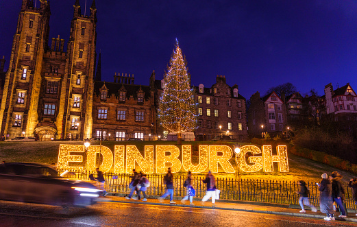 Pedestrians walking past a large illuminated sign on the Mound in central Edinburgh at night, with the Church of Scotland's General Assembly Hall building behind the Christmas tree.