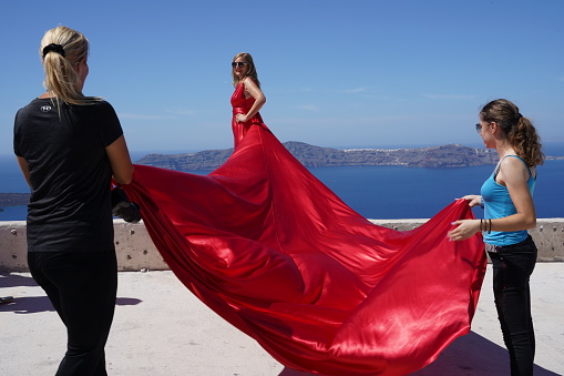 Santorin, Greece, 09 14 2023: Two assistants are holding the model's train of a red dress. The model is standing on a wall and behind her is the sea and an island.