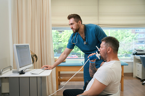 Adult man with nose clip breathing into mouthpiece of spirometer while doctor looking at computer screen