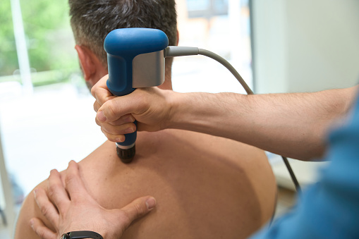 Cropped photo of physiotherapist using extracorporeal shockwave therapy device for upper back pain relief in adult man