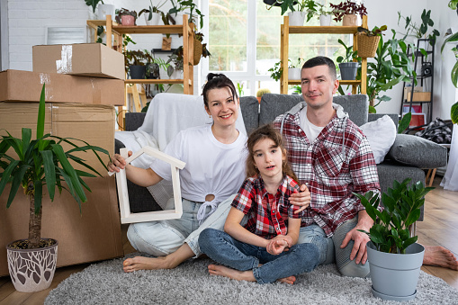 Moved family with a child and home plant are enjoying a new home, sitting on the sofa and holding the roof near boxes with things. Mortgage, insurance, buying and moving into your housing