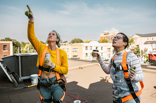 Two Female artists  painting roof top mural. They are dressed in casual work clothes. Exterior of old building in the city of Toronto, Canada..