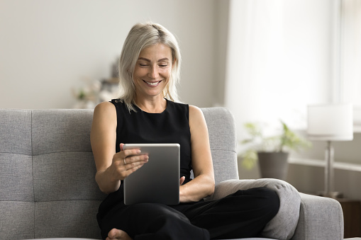 Cheerful pretty senior woman holding tablet computer, sitting on home couch, talking on video call, using e-commerce app for shopping, software for remote freelance job task, smiling, laughing